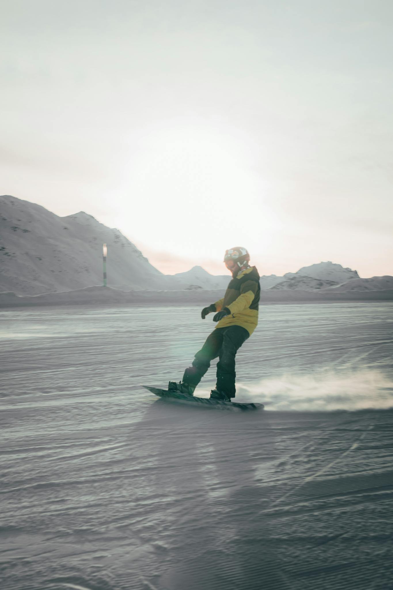 Snowboarder gliding down snowy slopes with mountains in the background under bright sunlight.