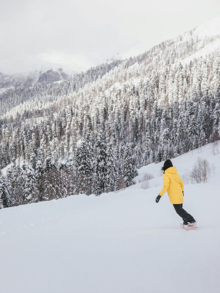 Snowboarder in yellow attire descends a snowy mountain slope surrounded by pine trees.